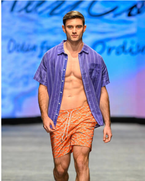 Model in an open purple shirt and orange shorts on the runway