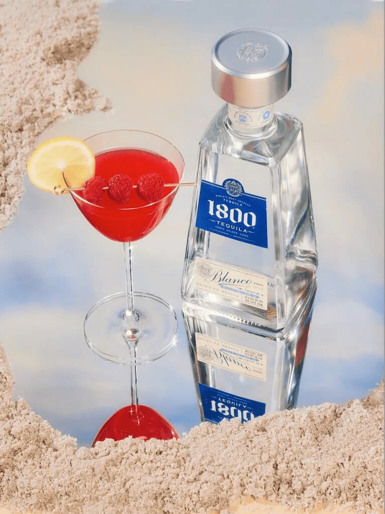 Tequila bottle and red cocktail on sand